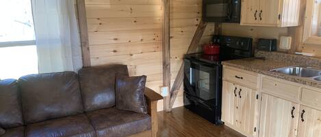 Interior acogedor de una cabaña con piso de madera, un sofá de cuero marrón, una zona de cocina con gabinetes de madera, un microondas, una estufa y una olla roja.
