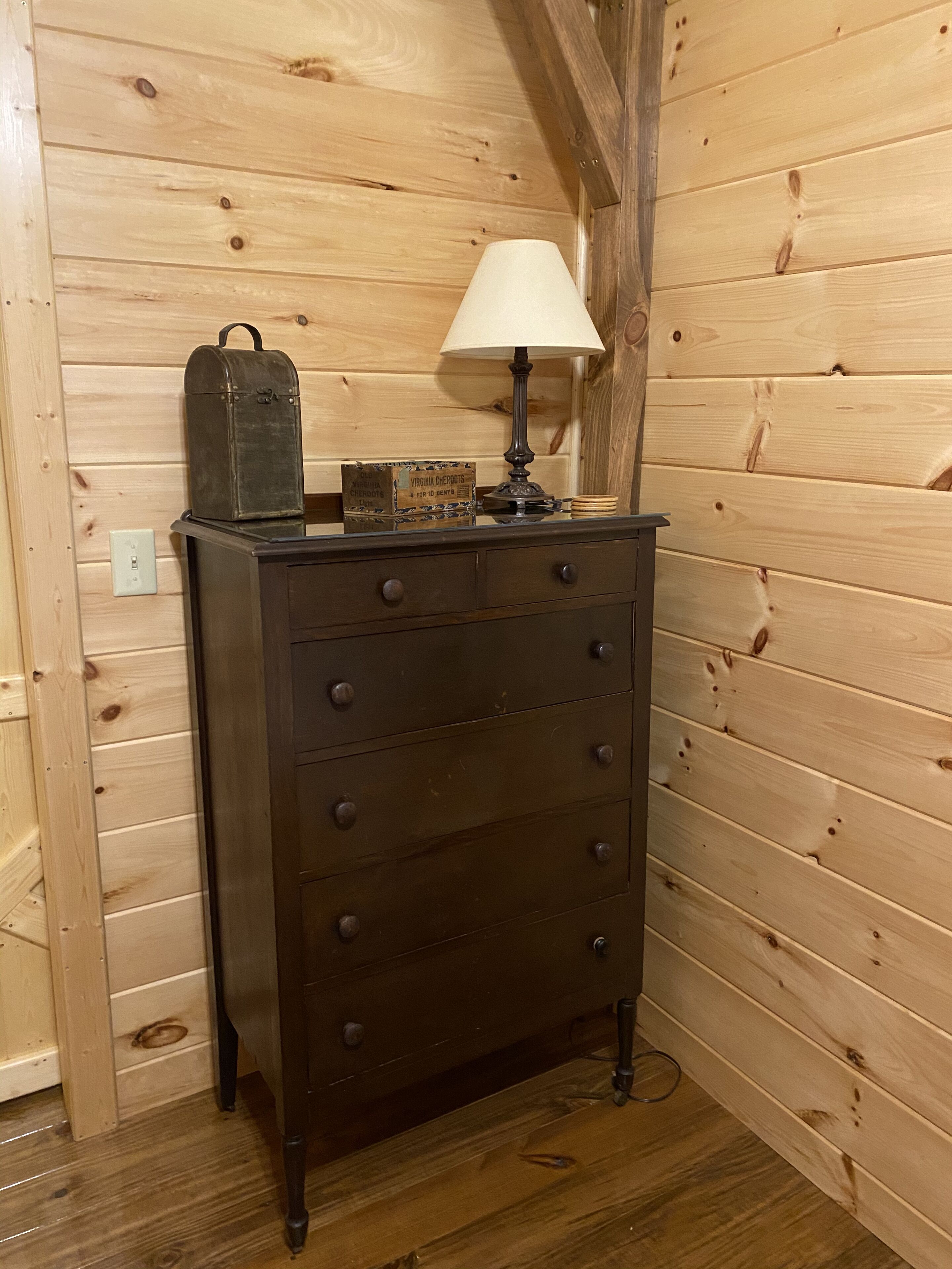 Un aparador de madera con cajones, una lámpara y una maleta antigua en una habitación con paredes y suelo de madera.
