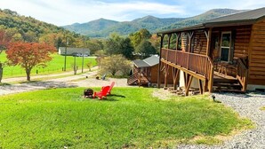 A log cabin with a porch and a covered deck, surrounded by a grassy yard and a mountain backdrop.