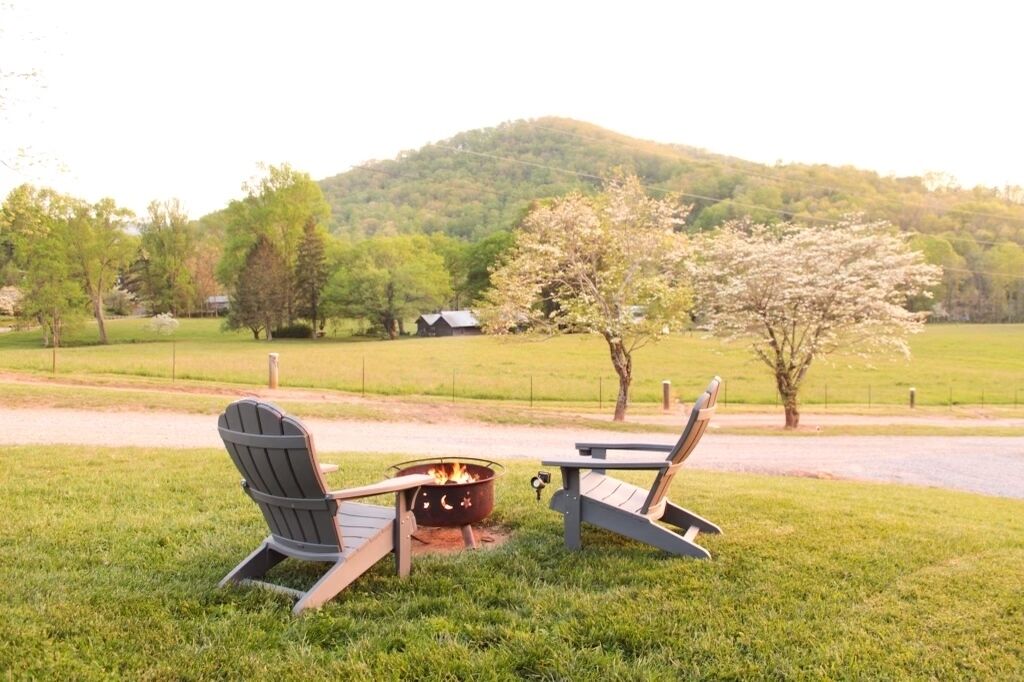 A campsite with two Adirondack chairs, a fire pit, and a view of a hill with trees and a house in the background.