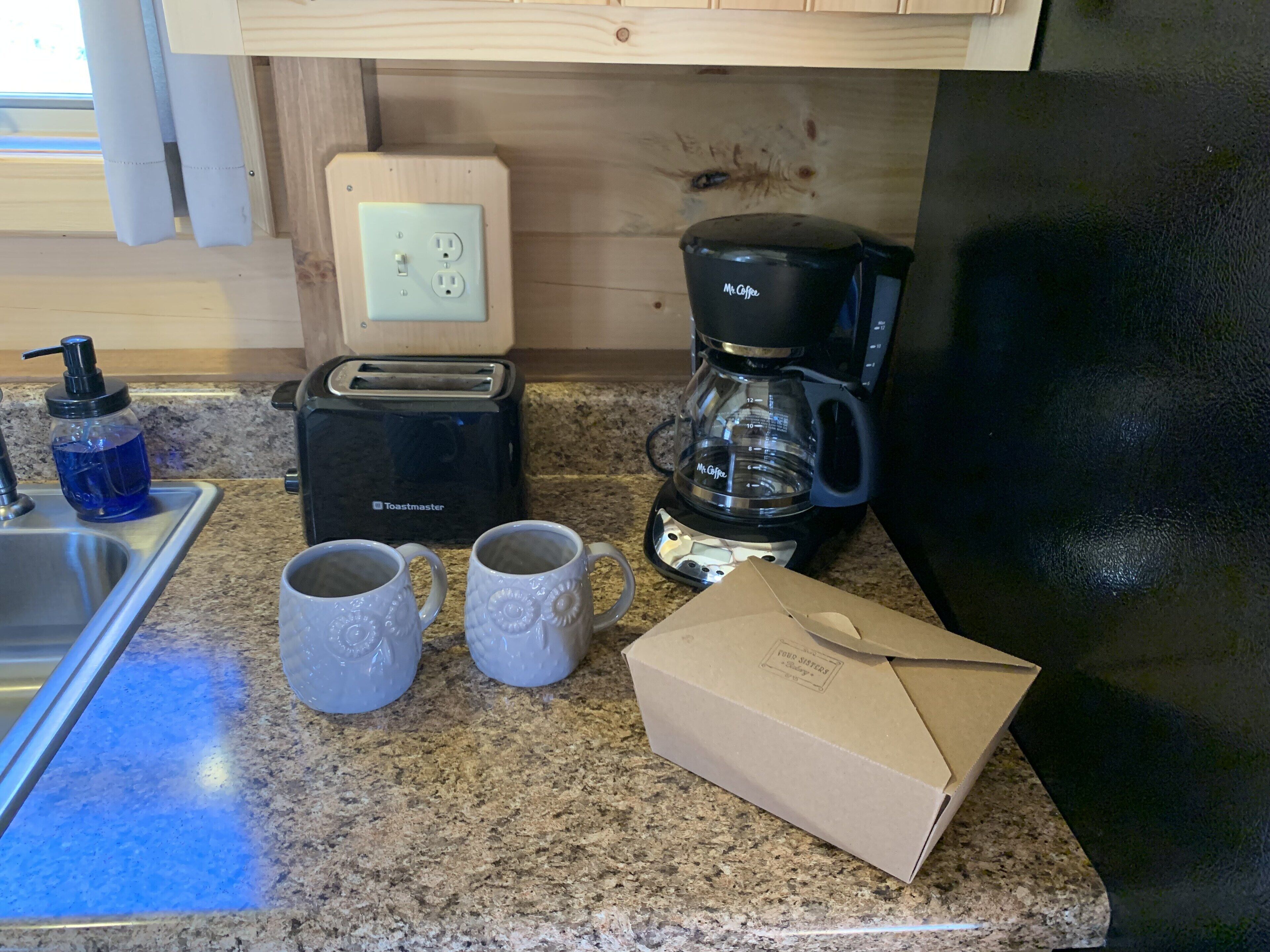 Una cocina con un tostador, cafetera, dos tazas, dispensador de jabón y una caja sobre la encimera.