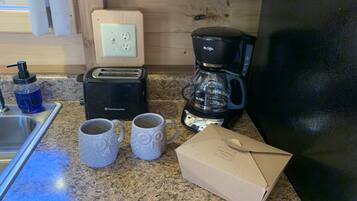 Una cocina con un tostador, cafetera, dos tazas, dispensador de jabón y una caja sobre la encimera.