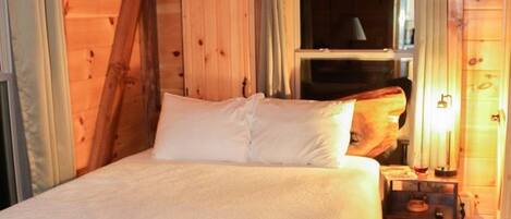 A wooden cabin bedroom with a bed, curtains, a nightstand, and a window.