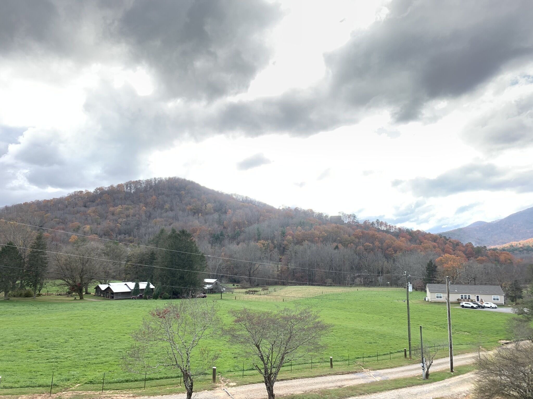 A rural landscape with a green field, a dirt road, and a hill with trees in autumn colors.