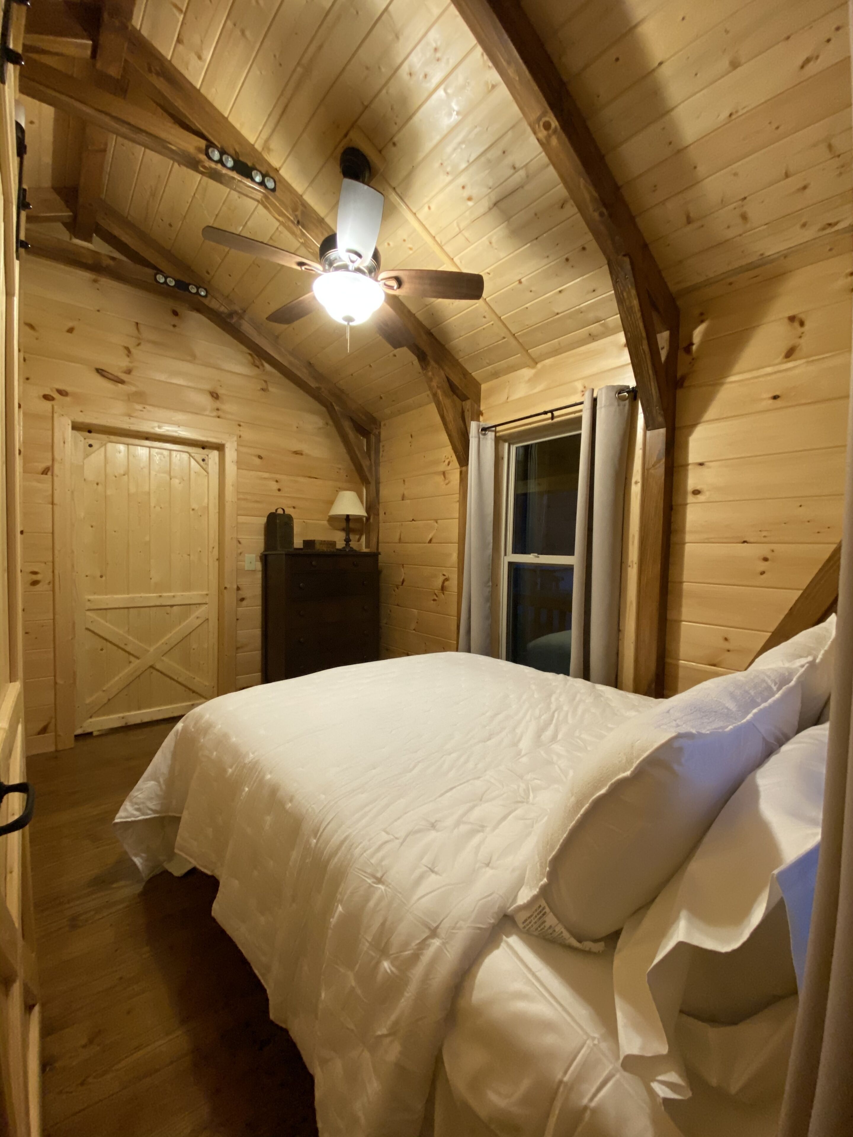 Dormitorio de cabaña de madera con cama, ventilador de techo, ventana y puerta.
