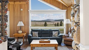 A cozy living room with a large window, wooden walls, a leather sofa, a coffee table, and a stone wall. - Pet-Friendly Retreat | Hot Tub, AC, Rec Center Access | Near Lake & Trails (Truckee)