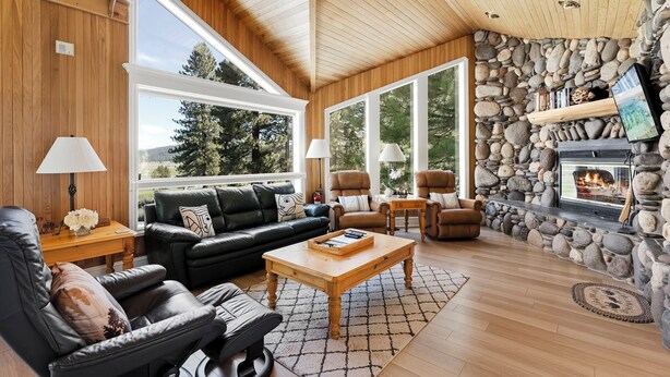 A cozy living room with a stone fireplace, leather furniture, and large windows offering a view of the outdoors. - Pet-Friendly Retreat | Hot Tub, AC, Rec Center Access | Near Lake & Trails (Truckee)