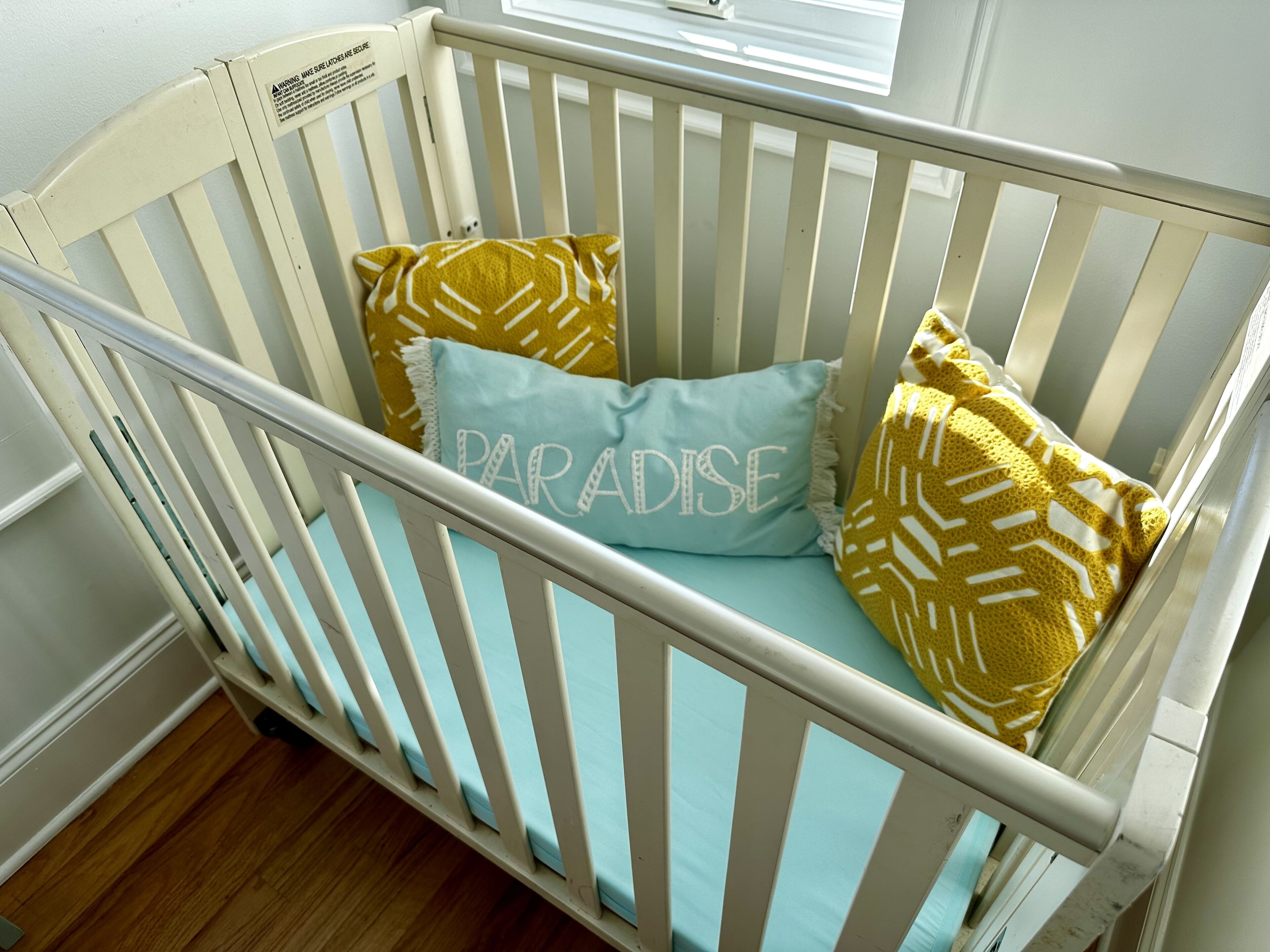A white crib with a light blue mattress and two pillows, one with a yellow pattern and the other with the word "PARADISE" embroidered.