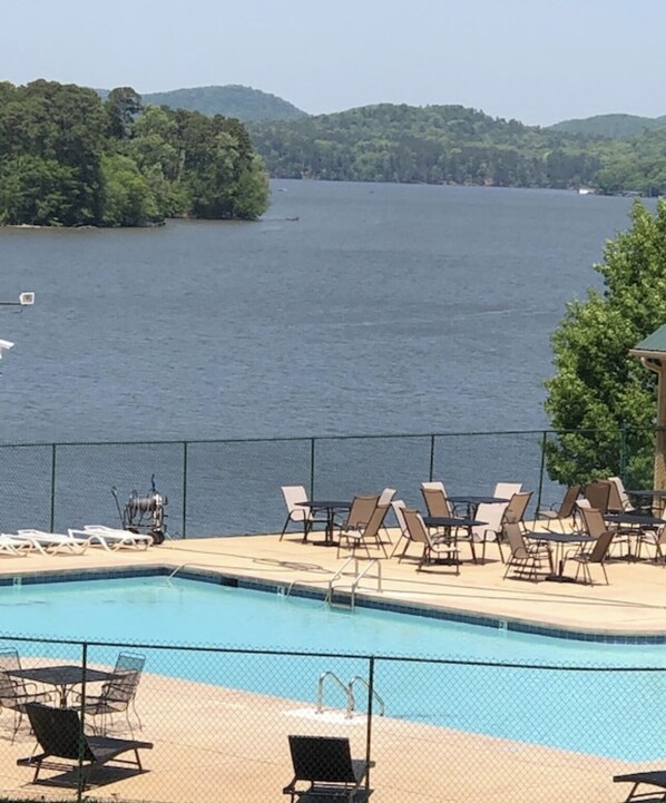 A pool area with lounge chairs and tables, surrounded by a fence, overlooking a lake with hills in the background.