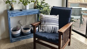 Un salon avec un fauteuil en bois, un téléviseur fixé au mur et une grande fenêtre.
