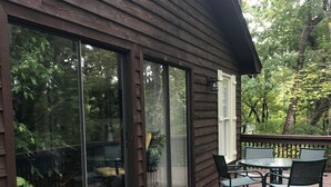 Une terrasse en bois avec un espace patio fermé par des baies vitrées, une table avec des chaises et une vue sur la forêt environnante.