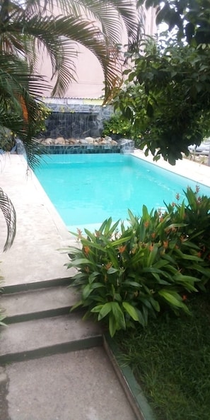 A swimming pool with a stone waterfall feature, surrounded by greenery and a concrete deck.