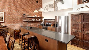 A kitchen with a brick wall, stainless steel appliances, wooden bar stools, and a large island. - Remodeled Penthouse in the heart of Downtown Basalt, historic 1900's Building  (Basalt)