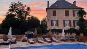 A two-story stone building with a swimming pool, outdoor seating, and a sunset sky.