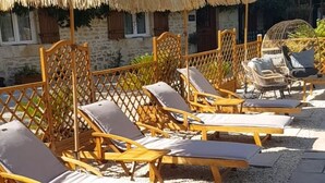 A row of wooden lounge chairs with cushions, arranged on a gravel patio with a thatched umbrella overhead.