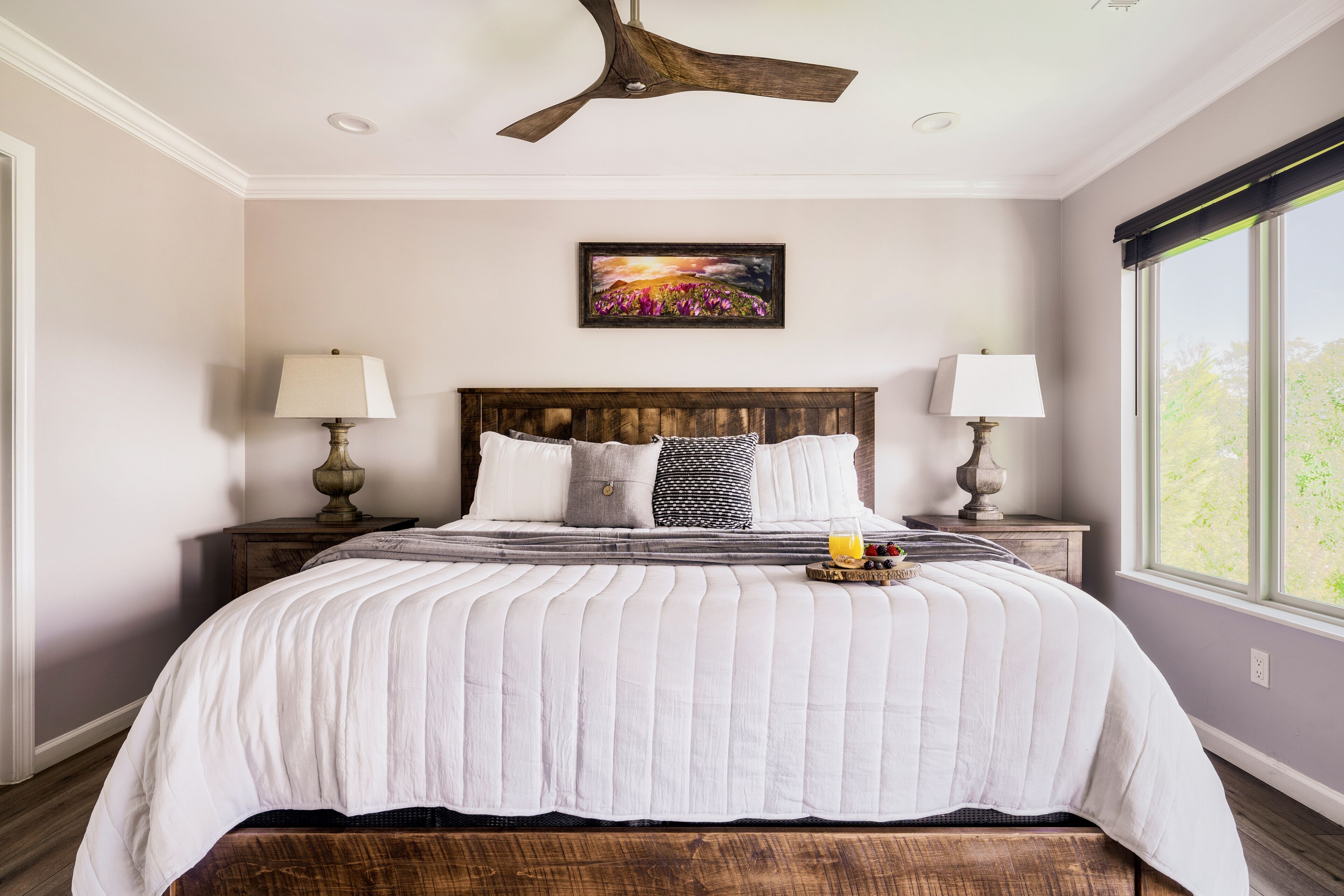 A bedroom with a large bed, two bedside tables with lamps, a ceiling fan, a framed picture on the wall, and a window with a view of greenery.