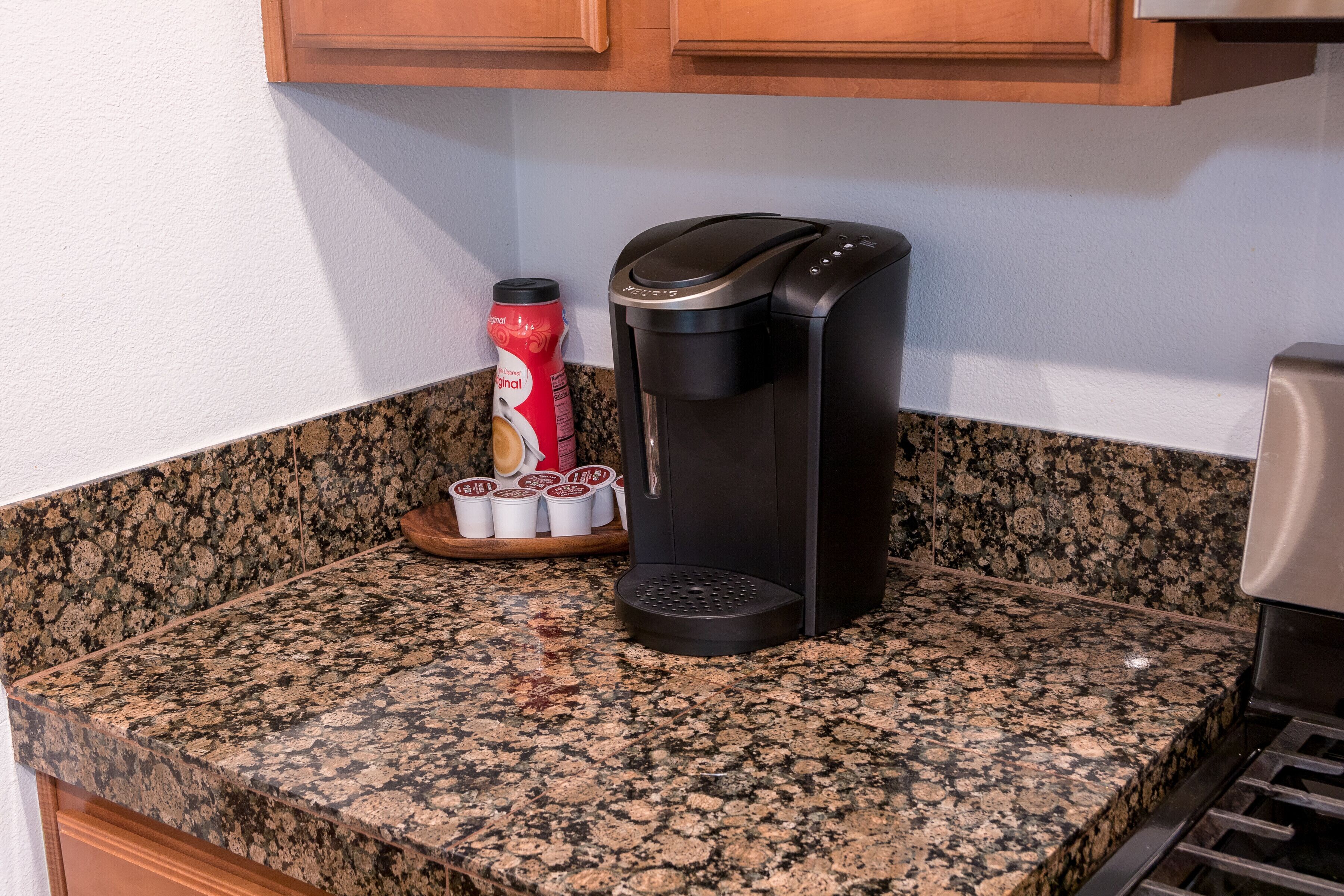 Une cuisine équipée d’une machine à café, d’une boîte de café et d’un plateau rempli de tasses à café, le tout posé sur un plan de travail en granit.