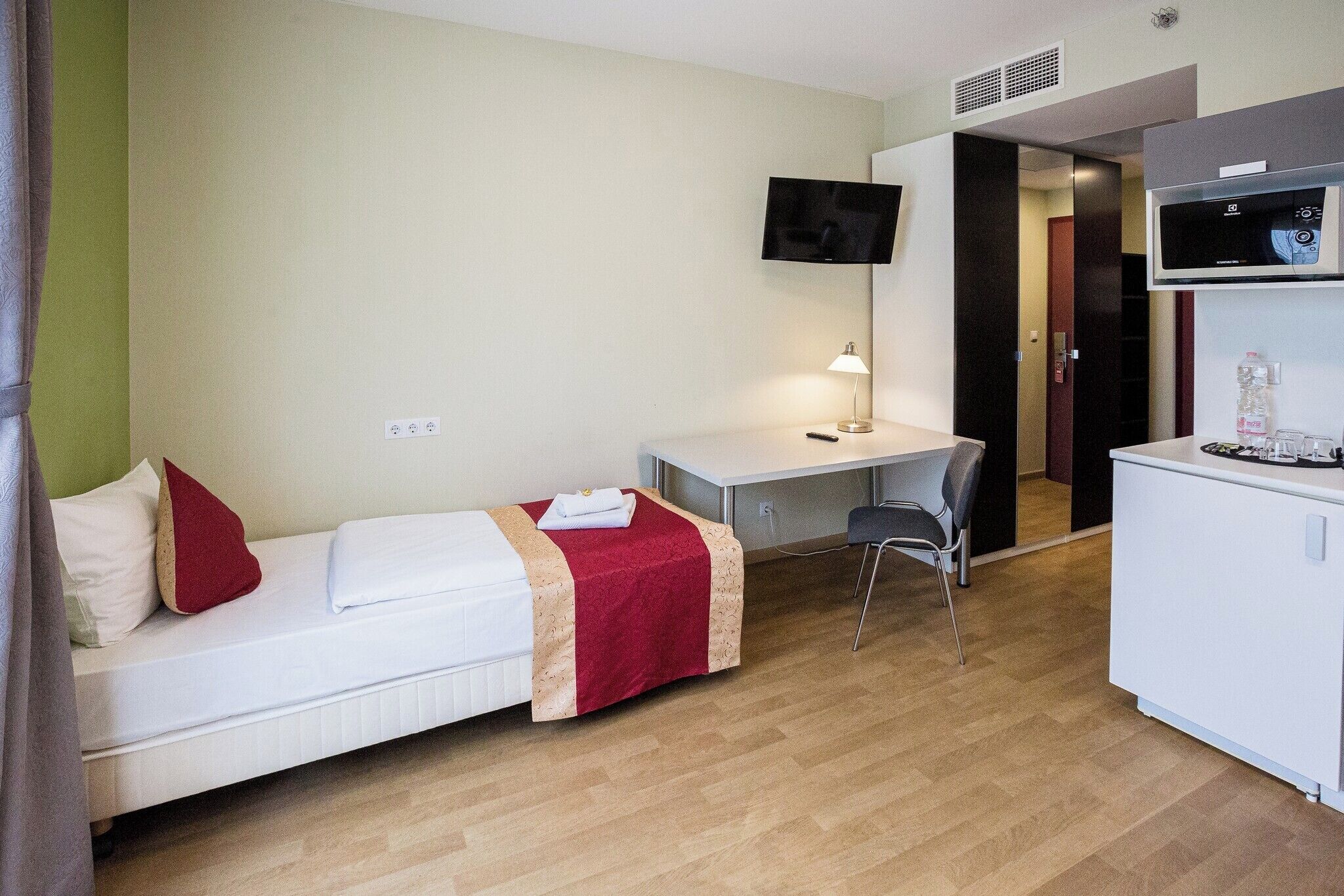 A hotel room with a bed, desk, chair, TV, and kitchenette.