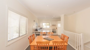 A dining area with a wooden table and chairs, a kitchen in the background, and a white railing along the wall. - Alaska - 8 Poley Cow Lane (Jindabyne)