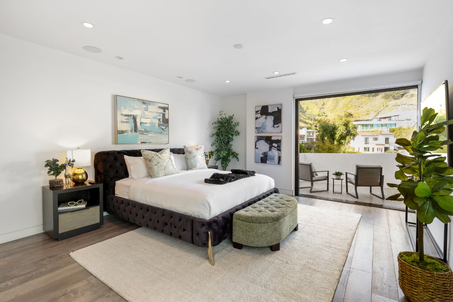 A modern bedroom with a large bed, a nightstand, a bedside table, a window with a view of buildings, and a balcony with chairs.