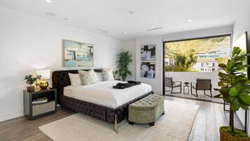 A modern bedroom with a large bed, a nightstand, a bedside table, a window with a view of buildings, and a balcony with chairs.