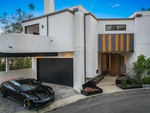 A modern two-story house with a garage, a black sports car parked in front, and a well-maintained garden.
