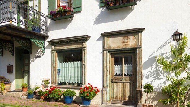 Ein Gebäude mit heller Fassade, grünen Fensterläden und einer Holztür. Vor den Fenstern stehen Blumentöpfe.