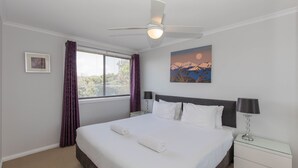 Un lit bien fait, avec du linge de lit blanc, un ventilateur de plafond, une table de chevet avec une lampe, une fenêtre avec des rideaux violets et un tableau représentant un coucher de soleil accroché au mur.