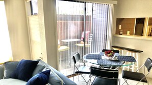 Una sala de estar con un sofá gris, una mesa de comedor de vidrio con sillas y un gran ventanal corredizo que da a un patio.