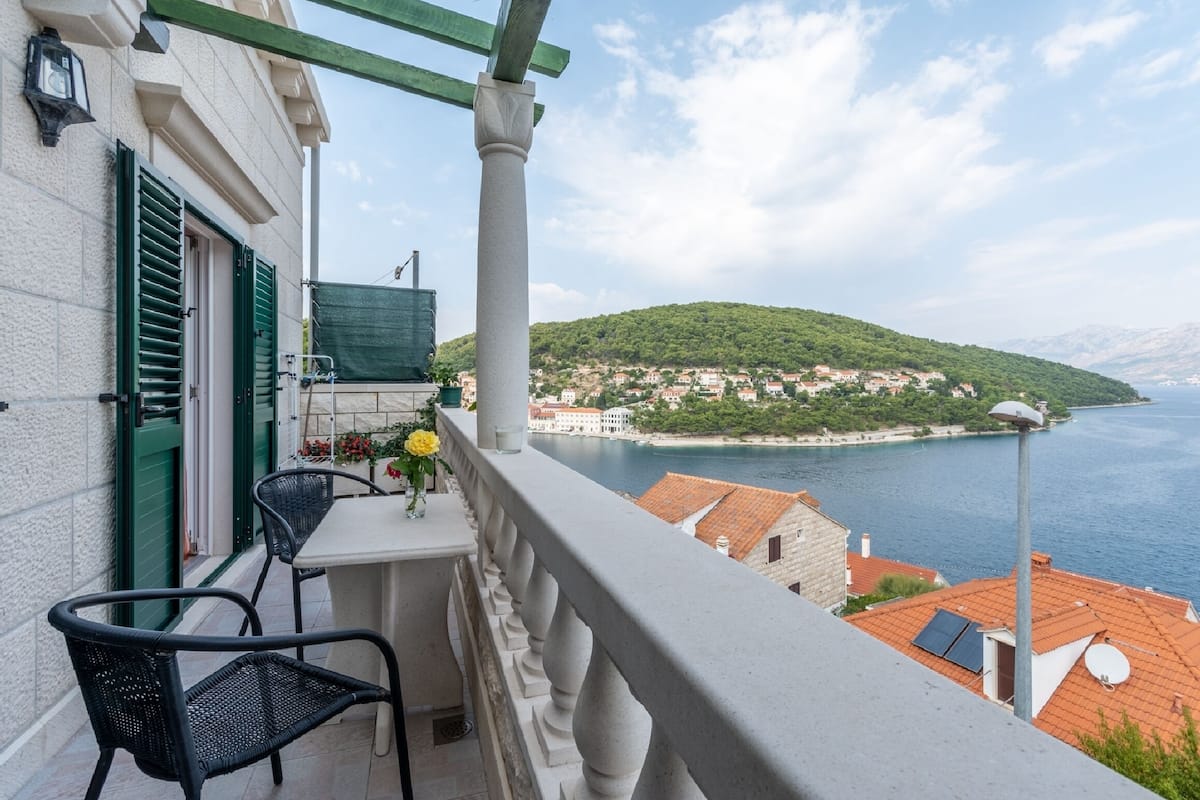 A balcony with a table and chairs, overlooking a coastal town and the sea.
