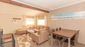 A cozy living room with a sectional sofa, a dining table with chairs, and a painting of a snowy landscape. - Charvel - 2/35 Gippsland Street (Jindabyne)