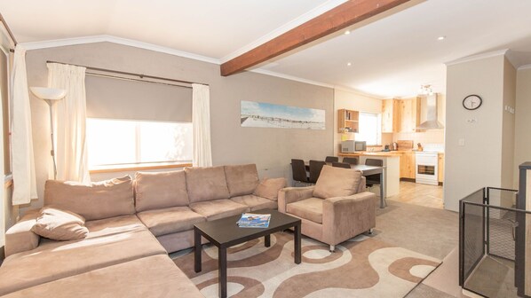 A living room with a sofa, armchair, and coffee table. There is a kitchen in the background with wooden cabinets and a dining area. - Charvel - 2/35 Gippsland Street (Jindabyne)