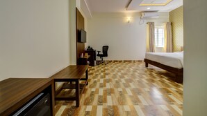 A hotel room with a wooden parquet floor, a bed, a desk with a chair, a TV mounted on the wall, and an air conditioning unit. - Hotel Royal Suites (Kolar)