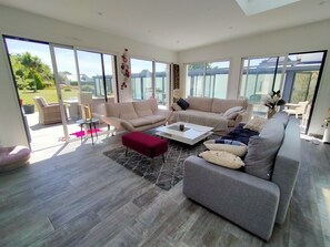 Ein geräumiges Wohnzimmer mit großen Schiebegläsern zum Balkon, ein beiges Sofa, ein graues Sofa und ein roter Hocker.
