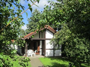 Et lille hus med et tag af fliser, omgivet af frodig vegetation og træer.