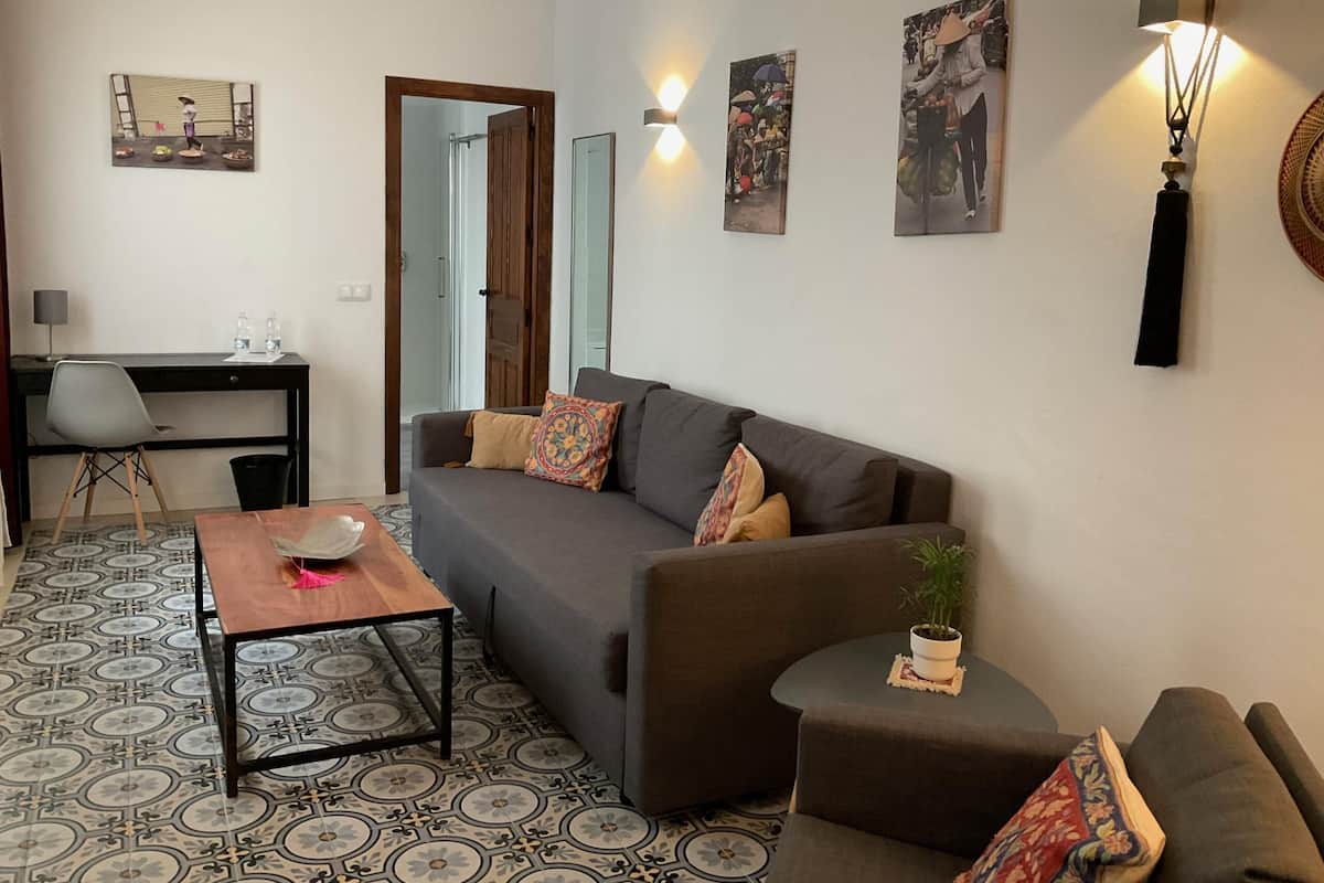 Un espacio habitable compacto con sofá, una mesita, un escritorio y una silla. Hay luces fijadas a la pared y un espejo.