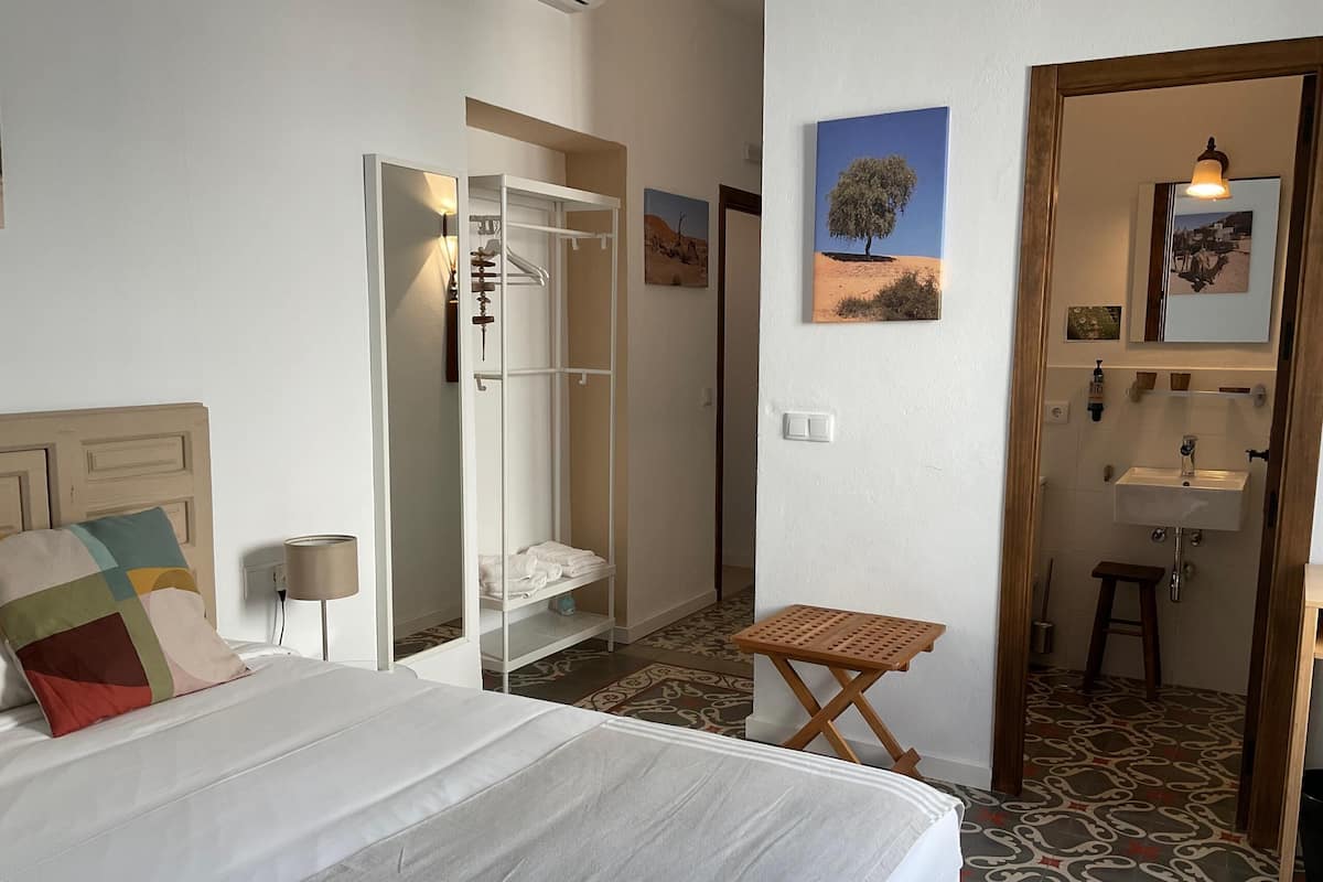 Una habitación de hotel con cama, vestidor, una mesita, espejo y baño con lavamanos y ducha.