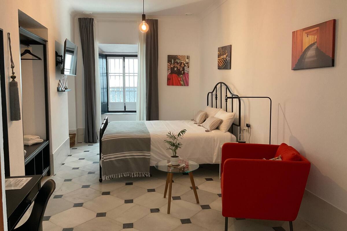 Un dormitorio con una cama, una silla roja, una mesita con una planta, un televisor en la pared y una ventana con cortinas.