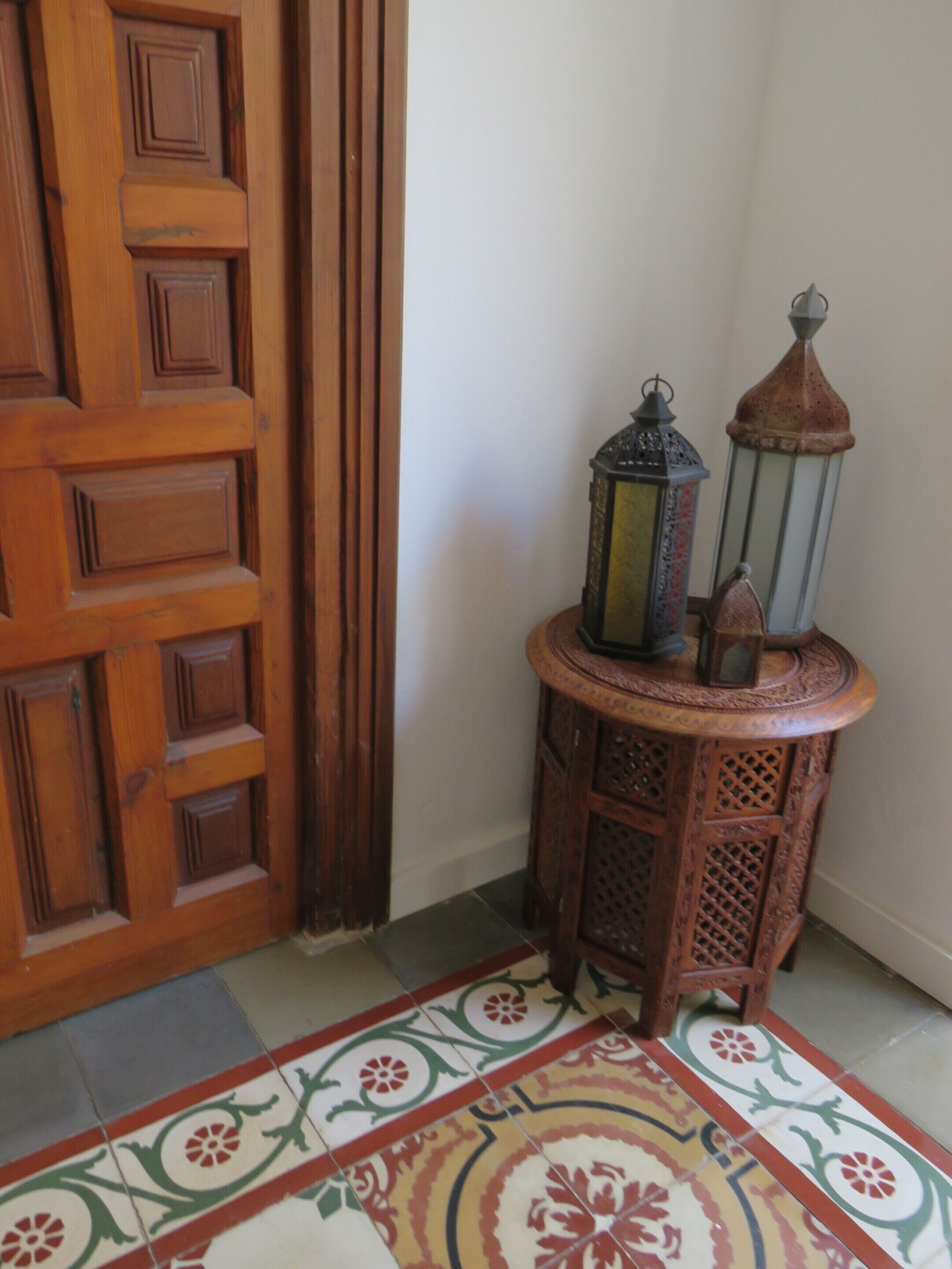 Une porte en bois ornée d’un panneau décoratif, une petite table en bois sur laquelle reposent deux lanternes, et un sol à motifs.