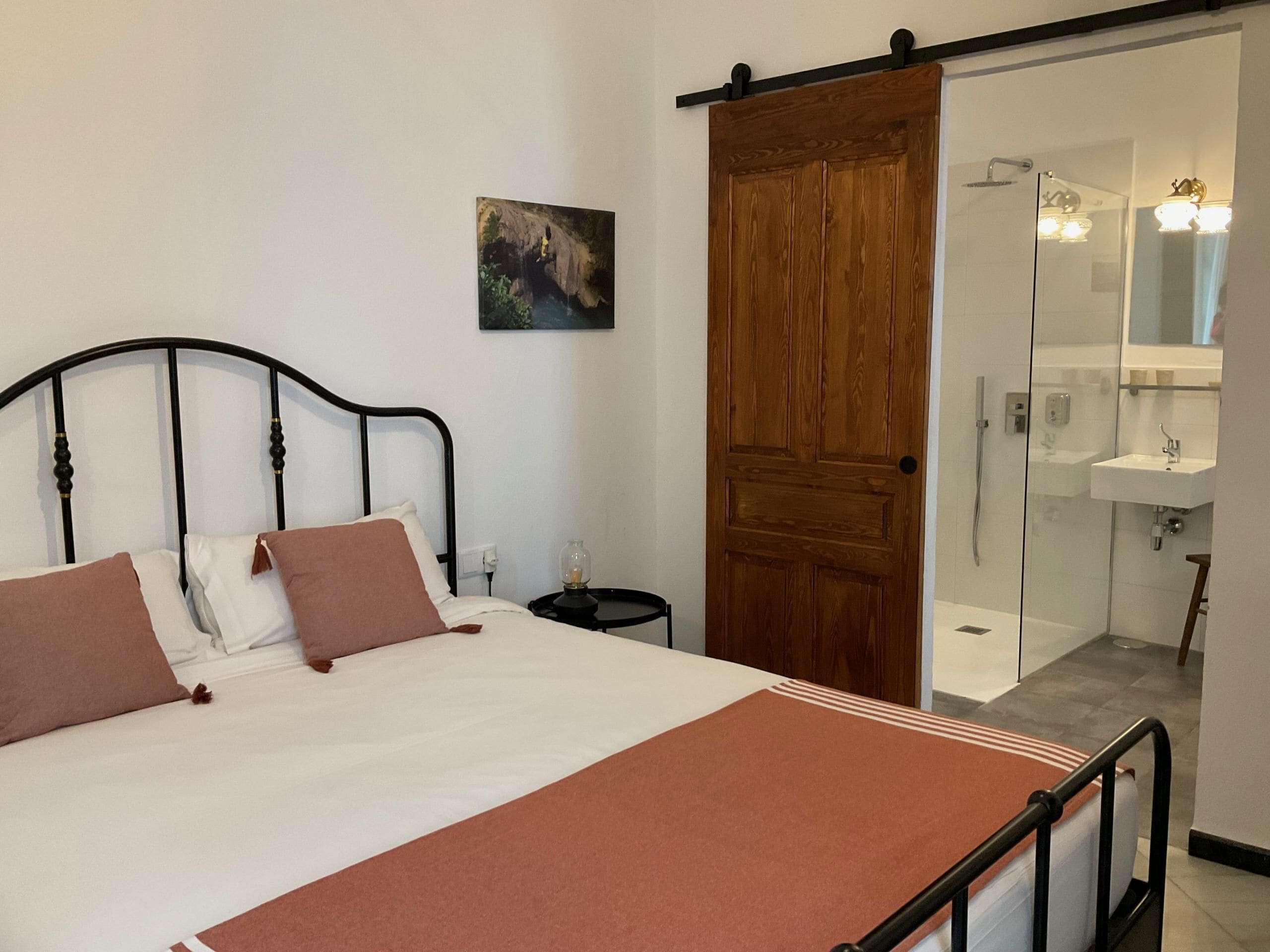 Un dormitorio con cama, puerta de madera, baño con ducha y una pequeña mesa con lámpara.