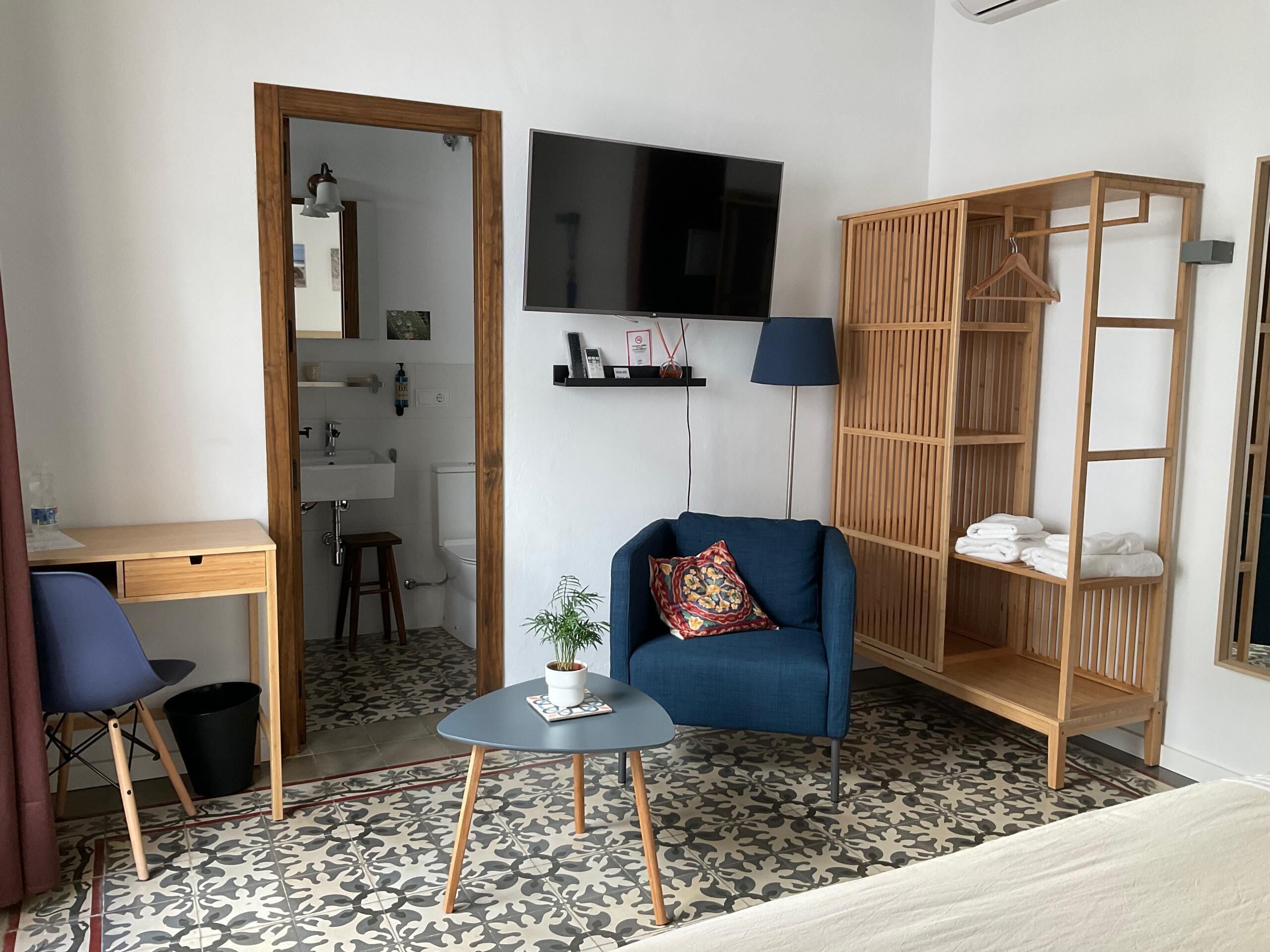 A compact room with a bed, a desk, a chair, a TV, a small table with a plant, and a wooden shelf with towels.