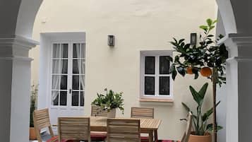 Un pati a l'aire lliure amb taula i cadires, plantes en testos i una paret blanca amb finestres.