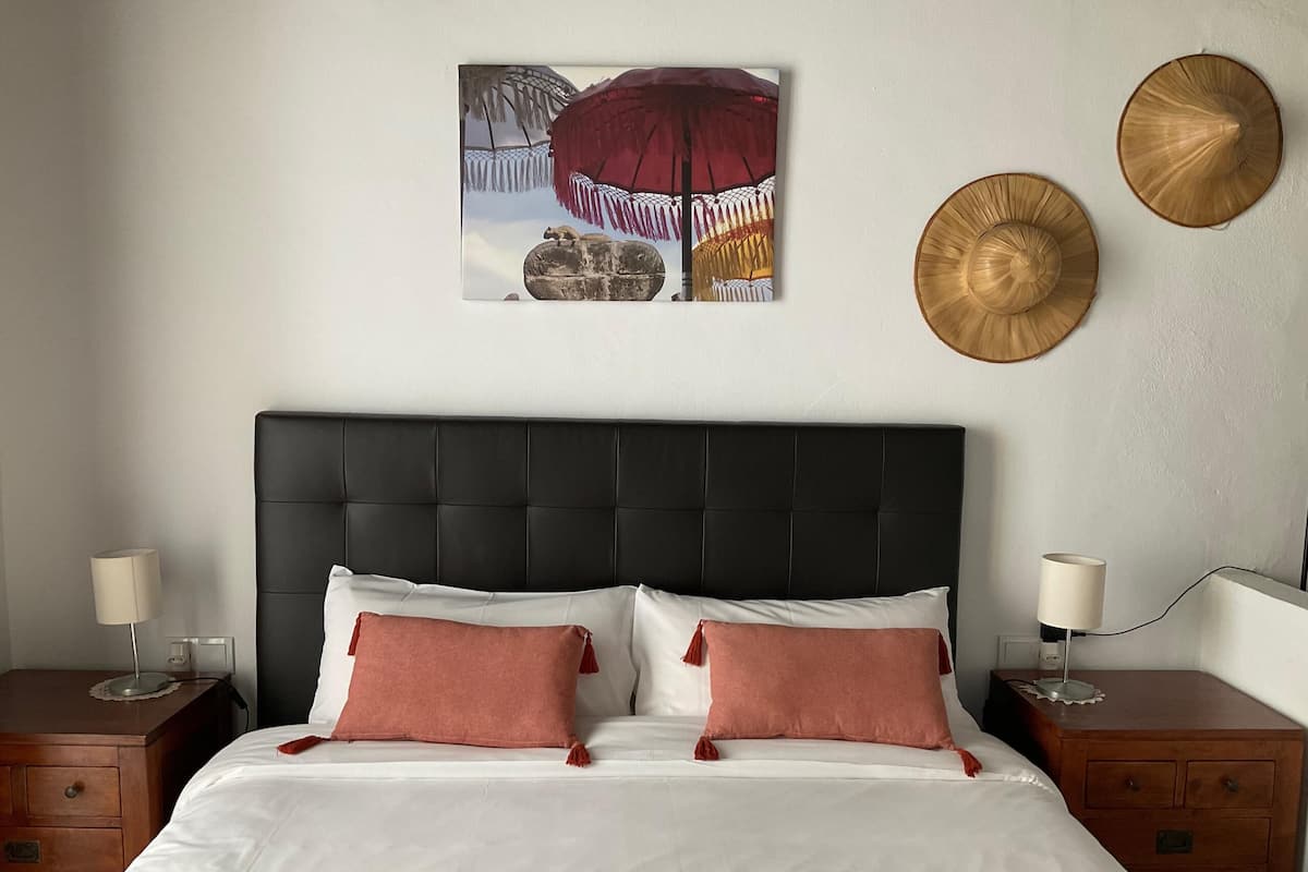 Un dormitorio con cabecera negra, ropa de cama blanca y dos almohadas rojas. Hay dos mesitas de noche con lámparas y una mesita auxiliar de madera.