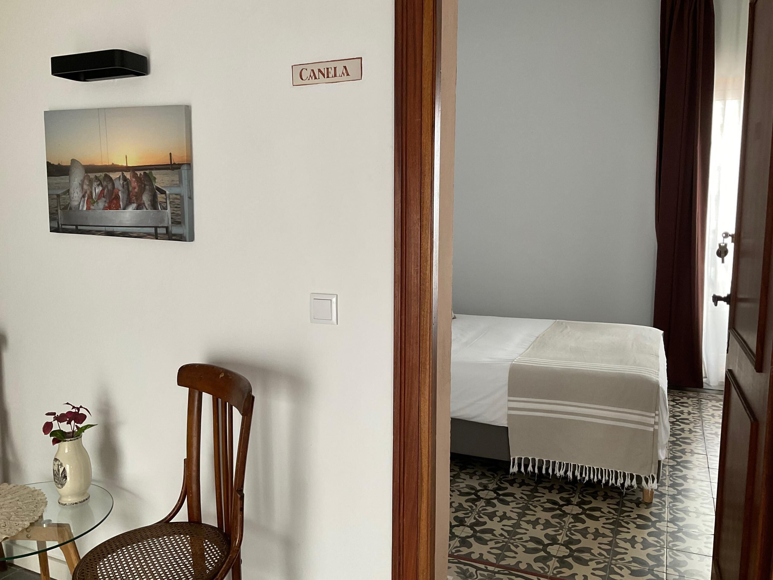 Una habitación con una cama, una silla, una mesita con un florero y un cuadro en la pared.