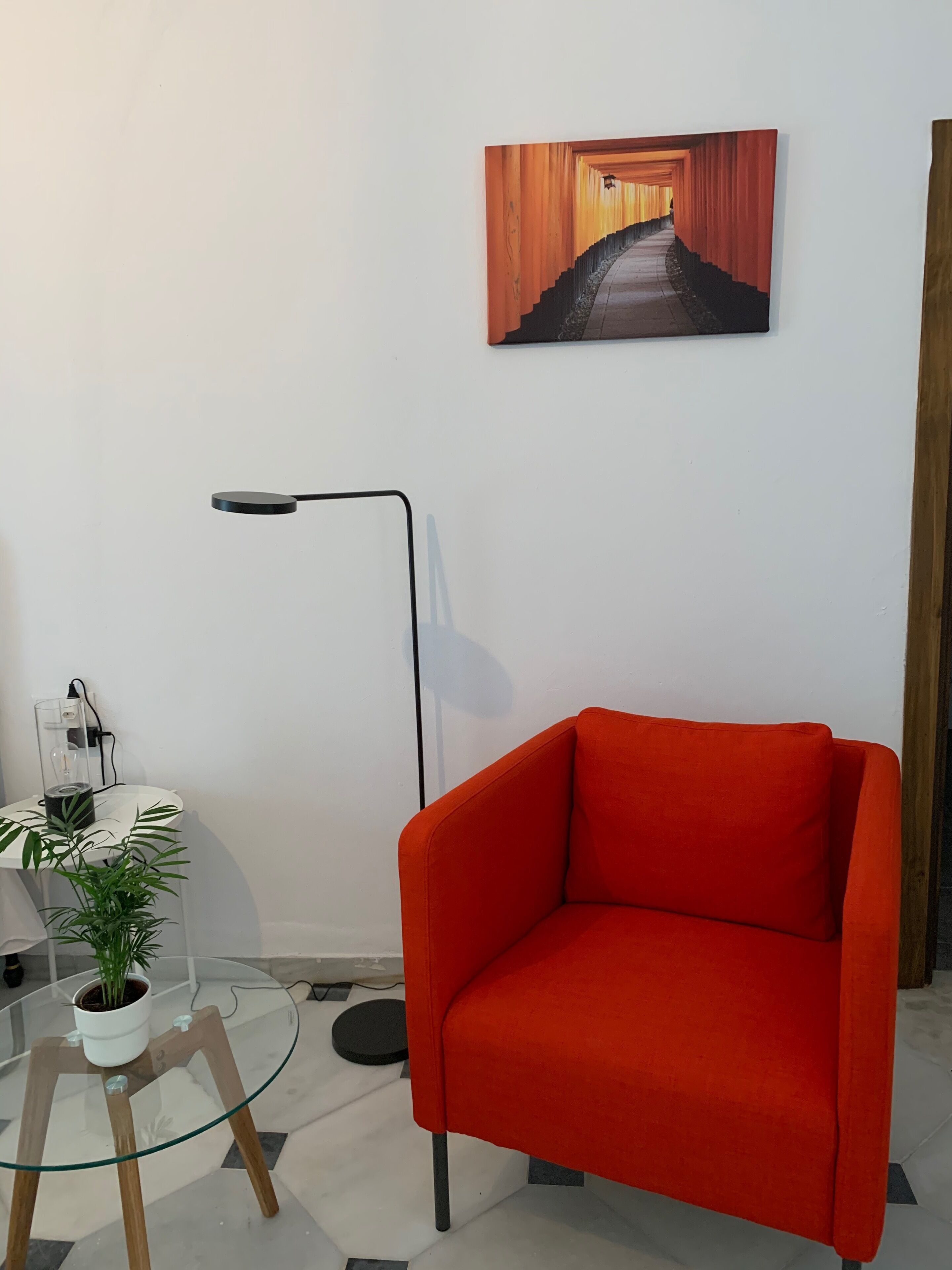 Una habitación moderna con un sillón rojo, una mesa de vidrio con una planta en maceta, una lámpara de pie y un cuadro en la pared.