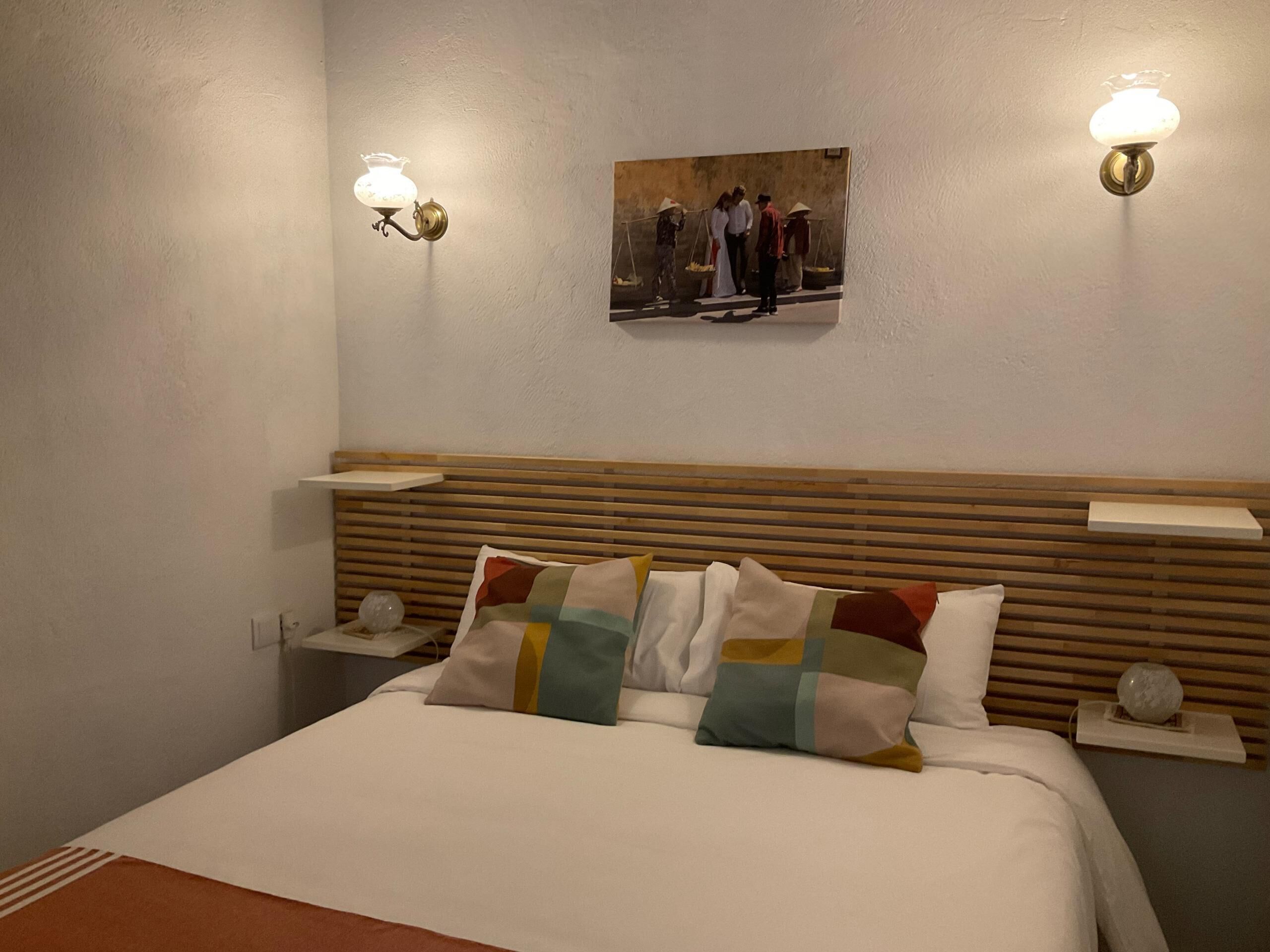 Habitación de hotel con cabecera de madera, cama con varios almohadones, dos lámparas de pared y un cuadro sobre la cama.