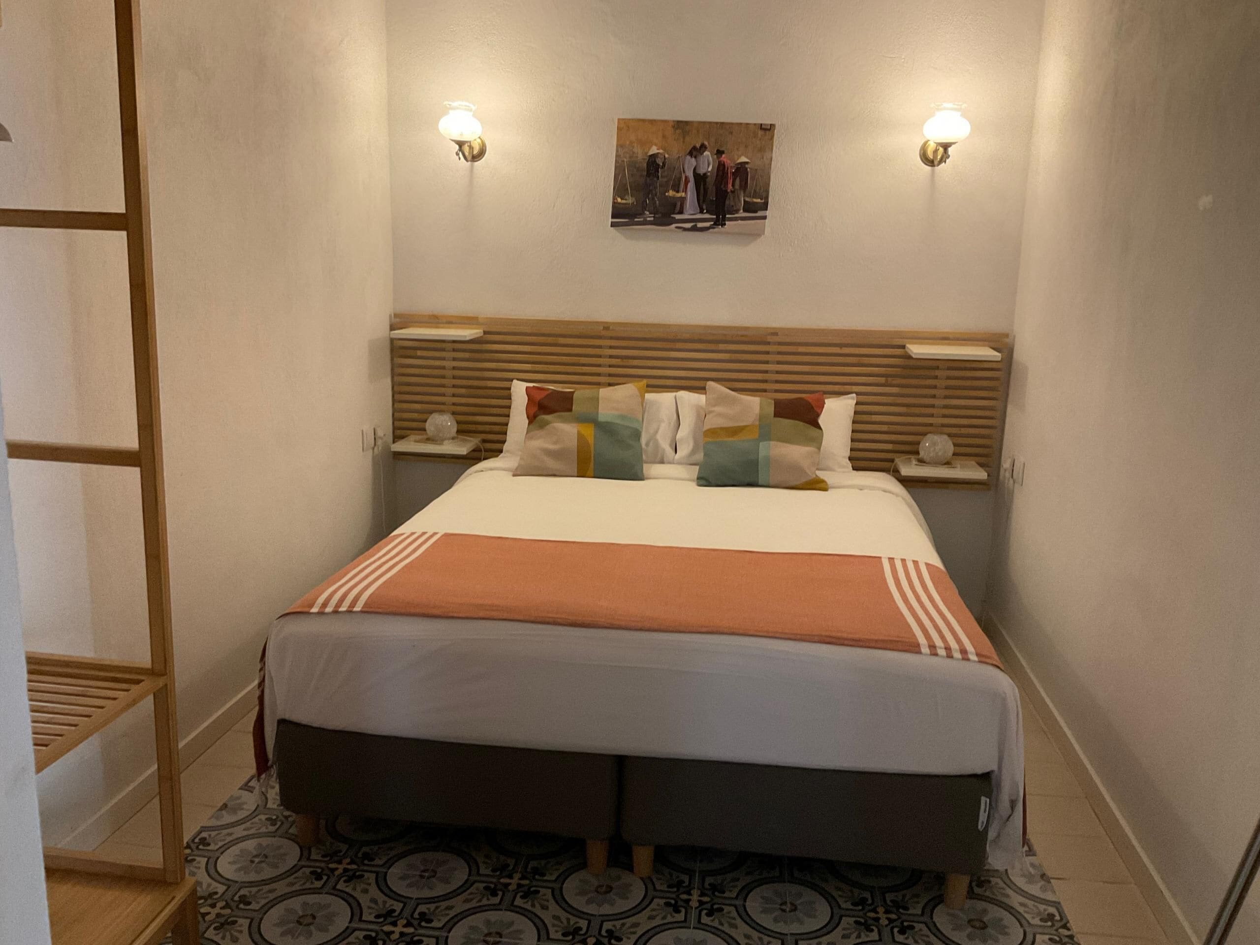 Une chambre d’hôtel comprenant un lit, deux tables de chevet, une petite table, un tableau accroché au mur et une tête de lit en bois.