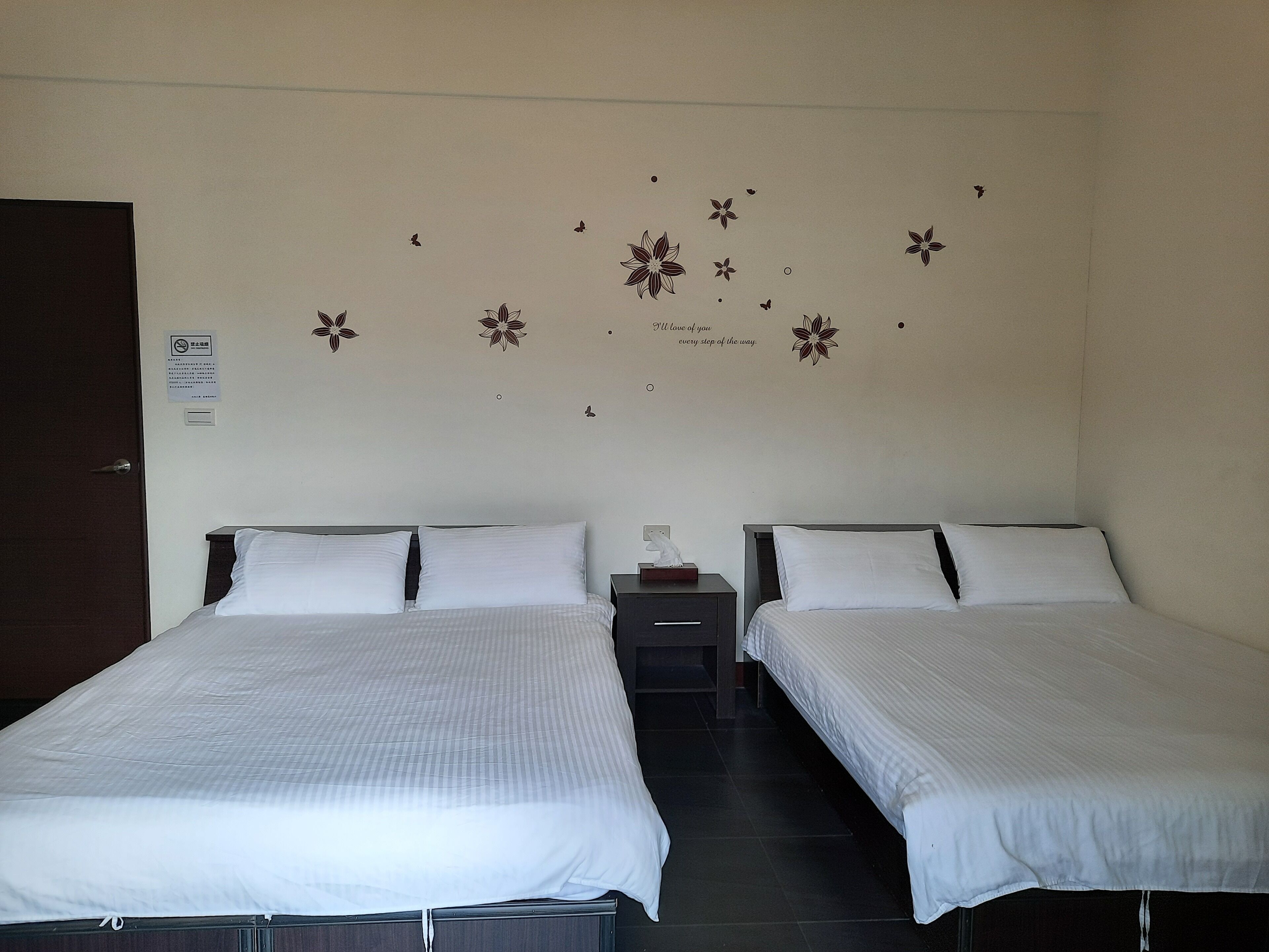 Habitación de hotel con dos camas individuales, una mesita de noche con lámpara y una pared decorada con motivos florales.