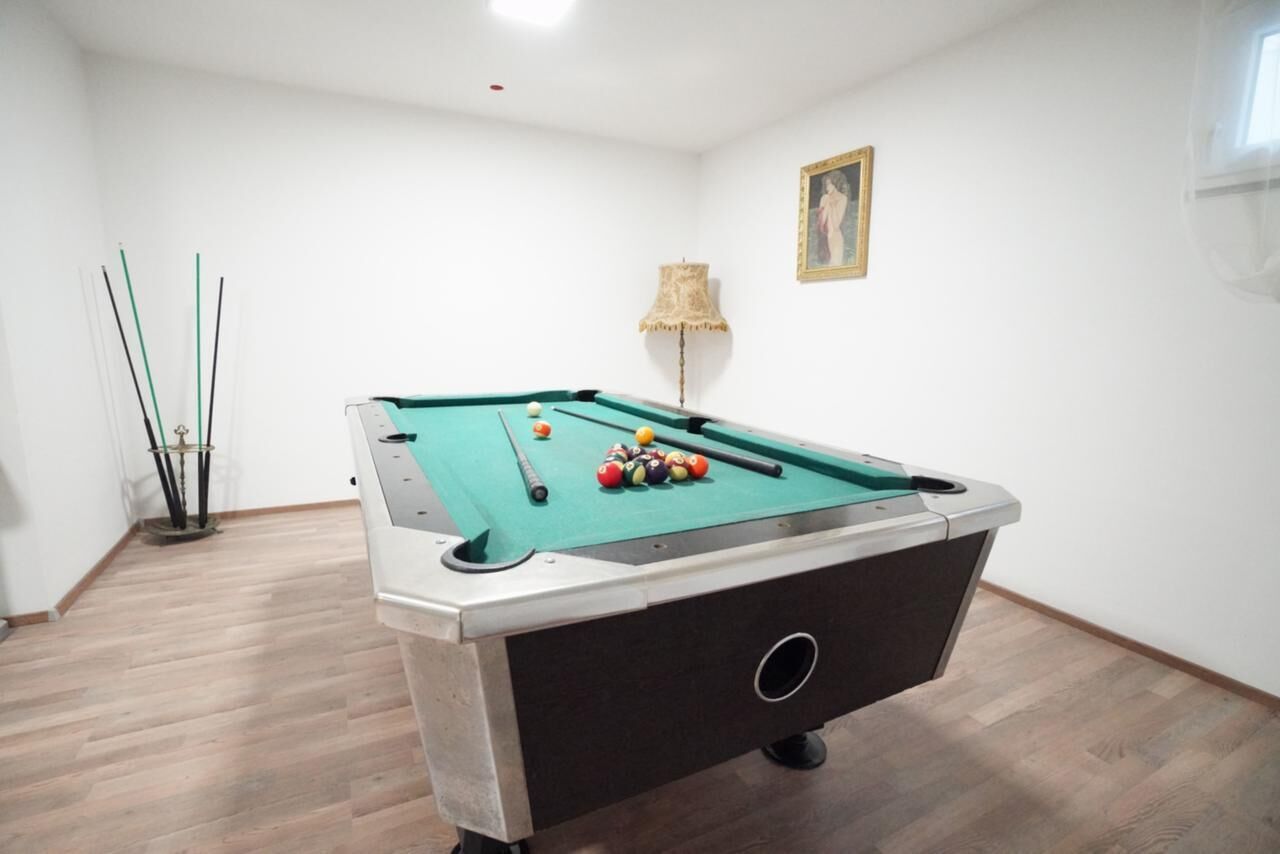 Une table de billard dans une pièce dont le sol est recouvert de parquet, avec une lampe et un tableau encadré au mur.