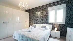 A bedroom with a large bed, white wardrobe, patterned wallpaper, a chandelier, and a window with blinds. - Villa Daniela (Solin)